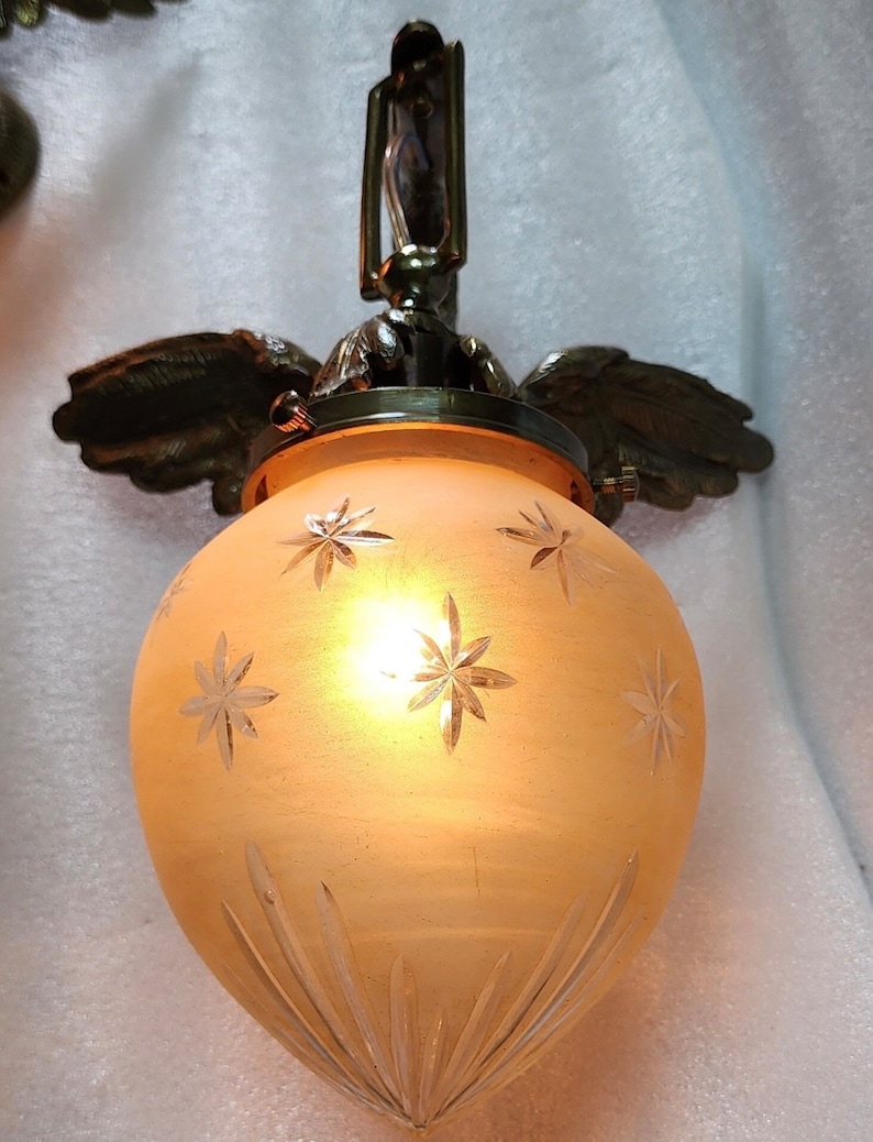 May include: A frosted glass pendant light fixture with a brass finish and a starburst pattern. The light fixture is suspended from a chain and has a decorative winged design.