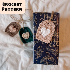 May include: Three crocheted heart-shaped gift tags with brown, green, and beige yarn. The tags are hanging from a gift wrapped in blue paper with a gold pattern and the words "Merry Christmas".