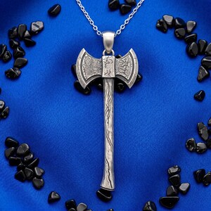 Può includere: Una collana con pendente a forma di ascia argentata con un design dettagliato, sospesa da una delicata catena. Il pendente a forma di ascia è posto su uno sfondo blu vibrante, circondato da pietre nere.
