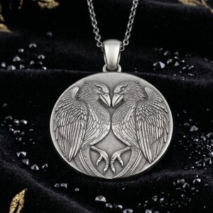 May include: A silver pendant necklace with a circular pendant depicting two facing birds with detailed wings. The pendant is suspended from a fine silver chain. The birds are intricately carved. The background is dark and textured.