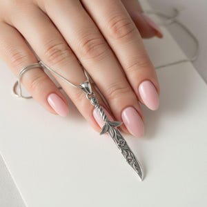 May include: A silver pendant necklace with a detailed dagger design and floral accents. The pendant hangs from a delicate chain. The image shows the necklace on a white surface.