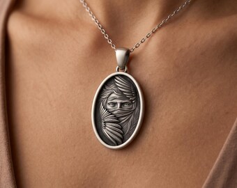 May include: An oval silver pendant necklace featuring a detailed relief carving of a face partially obscured by a veil and stylized feathers. The pendant hangs from a simple silver chain, showcasing a dark oxidized finish within the recessed areas.