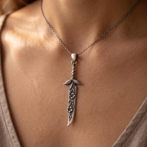 May include: A silver-toned necklace featuring a pendant shaped like a small sword or dagger. The blade is decorated with embossed floral and vine patterns, while the hilt has a twisted design. The pendant hangs from a simple, thin silver chain.