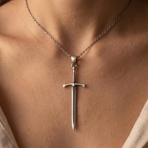 May include: A silver-toned metal necklace featuring a sword-shaped pendant. The pendant has a detailed hilt, a crossguard, and a straight blade. It hangs from a thin, delicate silver-coloured chain that rests against the collarbone area.