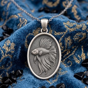 Puede incluir: Collar con colgante ovalado plateado con un relieve detallado de un pez betta. El colgante cuelga de una delicada cadena. El fondo es una tela azul texturizada con detalles dorados y piedras negras dispersas.