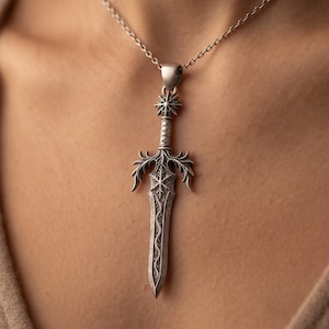 May include: A silver-toned metal pendant shaped like a sword, featuring intricate engraved scrollwork and a snowflake-like emblem at the crossguard. The pendant hangs from a delicate silver-toned chain, showcasing a pointed blade and detailed hilt.