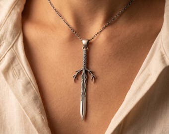 May include: A silver-toned metal pendant shaped like a long sword with a braided handle and curved, antler-like crossguard details. The pendant hangs from a thin, matching silver-toned metal chain, resting against a light-colored collared shirt.