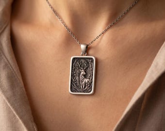 May include: A rectangular silver-toned pendant featuring a detailed embossed relief of a stag standing within a forest setting. The pendant hangs from a thin silver-toned chain, showcasing a polished frame and an oxidized finish on the central design.