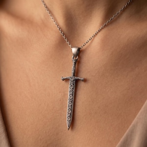 Op de afbeelding: Een zilverkleurige metalen hanger in de vorm van een zwaard, voorzien van gedetailleerde bloemengravures op het lemmet en het gevest. De zwaardhanger hangt aan een fijne zilverkleurige ketting tegen een neutraal gekleurde stoffen achtergrond.