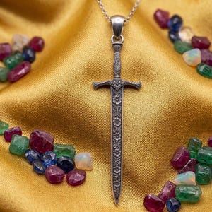 Può includere: Un ciondolo a forma di spada d'argento con dettagli floreali è appeso a una catena d'argento. Il ciondolo è circondato da pietre preziose colorate, tra cui rubini, smeraldi e opali, su uno sfondo di raso dorato. Le pietre preziose sono di varie forme e dimensioni.
