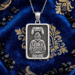 May include: Silver pendant necklace with a rectangular pendant featuring a gamer design. The pendant shows a person in a hoodie with the word "GAMER" and a game controller. The necklace is set against a blue background with gold floral patterns.