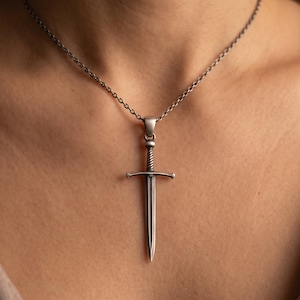 May include: A silver-toned metal necklace featuring a sword-shaped pendant with a twisted handle design and a pointed blade. The pendant hangs from a thin, matching silver-toned chain that rests against the skin, showcasing a metallic finish.