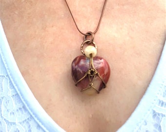 May include: A heart-shaped pendant necklace with a red, white, and brown stone set in a copper wire cage. The pendant is suspended from a brown chain.