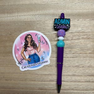 May include: A purple pen with a black tip and decorative beads. The pen has a charm that reads "ADMIN team" in teal and black. A round sticker with a woman holding a drink is next to the pen. The sticker has the text "CreationsbyStephhh".