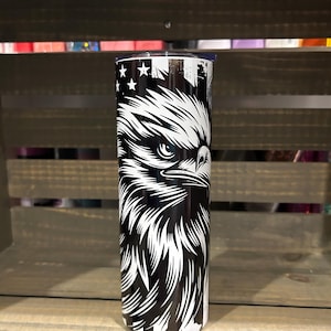 May include: A stainless steel tumbler featuring a detailed black and white illustration of an eagle's head. The design incorporates elements of the American flag, including stars and stripes. The tumbler is cylindrical and appears to be insulated.