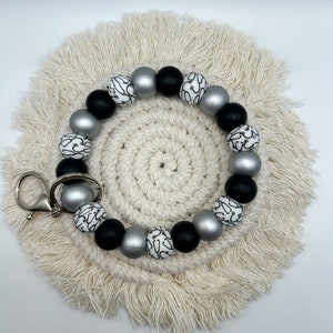 May include: A beaded keychain wristlet featuring a mix of black, silver, and white beads. The white beads have a black, abstract design. The wristlet is attached to a silver-colored clasp. The wristlet is resting on a cream-colored, woven coaster.