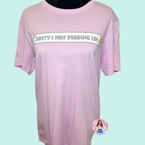 May include: A light pink t-shirt with the text "sorry I was reading lol" in a white banner. The shirt is displayed on a black mannequin. A small flower and a smiling face are on the banner.