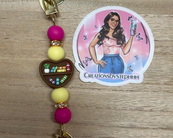 May include: A gold-colored keychain with a heart-shaped clasp and charm. The keychain features a series of beads in pink, yellow, and clear with rhinestone accents. A brown heart-shaped charm with a bookshelf design is also included. A sticker with the text "CreationsbyStephhh" is also visible.