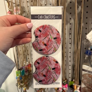 May include: Two pink and white car coasters with a pattern of scissors, nail files, and lipstick. The coasters are packaged together and have the text "...CAR COASTERS..." and "@CREATIVE_STEPNHH" printed on the packaging.