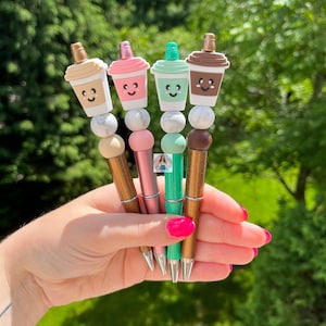 May include: Four colorful pens with cute coffee cup toppers. Each pen has a different color cup and a marble-like bead. The pens are held in a hand.