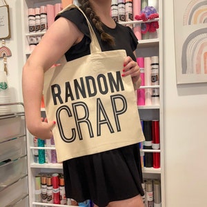 May include: A white canvas tote bag with black text that reads "RANDOM CRAP". The bag is being held by a person wearing a black shirt.