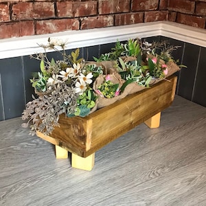 May include: A wooden planter box with four legs, filled with artificial plants and flowers. The planter is painted a dark brown colour and has a rustic look. The planter is sitting on a light grey wood surface.