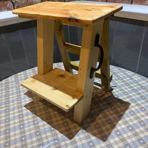 May include: A wooden step stool with a top shelf and a lower shelf. The stool is made of natural wood and has a black metal handle.
