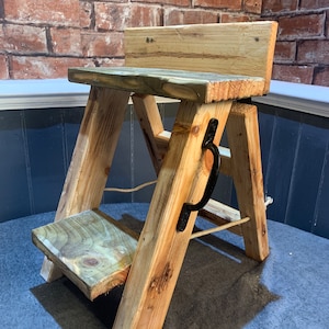 May include: A wooden step stool with a single step and a top shelf. The stool is made of reclaimed wood and has a rustic look. It has a black metal handle on the side.