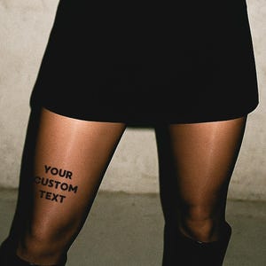 Può includere: Mini gonna nera e collant neri trasparenti. Il testo "YOUR CUSTOM TEXT" è stampato sulla coscia. L'outfit è completato da stivali neri. Lo sfondo è grigio neutro.