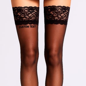 May include: Black thigh-high stockings with a delicate lace trim at the top. The stockings feature the text "CUSTOM TEXT" printed on the thigh. The sheer material creates a sleek and elegant look. These stockings are a stylish accessory.