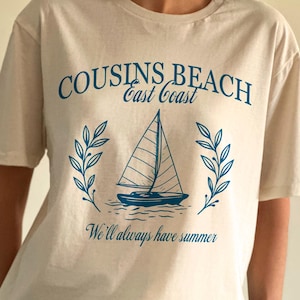 Könnte beinhalten: Beiges T-Shirt mit blauem Segelboot-Motiv, dem Text "Cousins Beach East Coast" und dem Satz "We'll always have summer". Das Shirt hat kurze Ärmel und einen Rundhalsausschnitt.