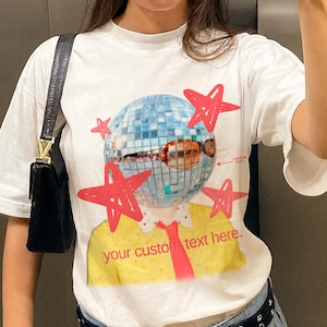 Puede incluir: Camiseta blanca con un diseño gráfico de una cabeza de bola de discoteca y estrellas rojas. El diseño incluye el texto "your custom text here." La camiseta tiene un cuello amarillo y una corbata roja.