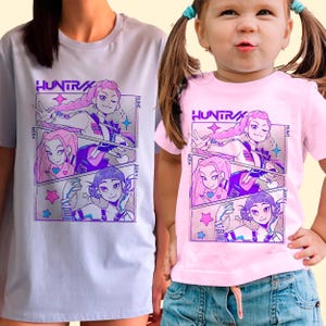 Puede incluir: Dos camisetas, una lila y otra rosa, con un gráfico de estilo cómic. El gráfico incluye la palabra "HUNTR" y tres paneles con ilustraciones de chicas estilo anime. Las camisetas son de manga corta.