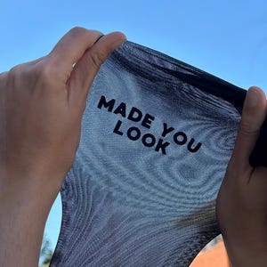 May include: Black sheer fabric held up against a blue sky. The text "MADE YOU LOOK" is printed in bold, white letters on the fabric. The fabric is being stretched between two hands.