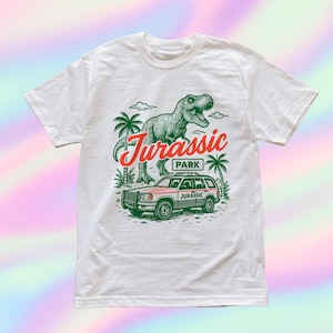 Pode incluir: Camiseta branca com uma ilustração de dinossauro verde e o texto "Jurassic Park" em vermelho e verde. O design inclui um SUV vermelho e verde e palmeiras, evocando um tema de filme retrô.