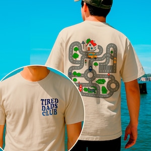 Puede incluir: Camiseta beige con un diseño de mapa de carreteras en la espalda, con una casa, autobús y coches. La parte delantera de la camiseta tiene el texto azul "TIRED DADS CLUB". La camiseta es de material suave.
