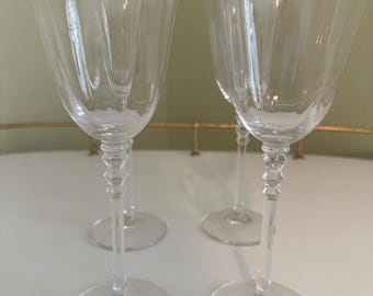 May include: Four clear wine glasses with a gold rim and a decorative stem. The glasses are arranged on a white surface, suggesting a setting for a special occasion or elegant dining.