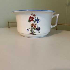 May include: White ceramic chamber pot with a blue rim and a handle. The pot is decorated with a floral design featuring red, blue, yellow, and purple flowers. The pot is sitting on a white surface.