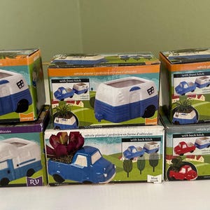 Puede incluir: Conjunto de seis macetas de cerámica con forma de vehículos antiguos. Las macetas son blancas con detalles azules y tienen una pequeña planta en cada una. Las macetas están etiquetadas con "con enganche para plantas" y "con enganche trasero".