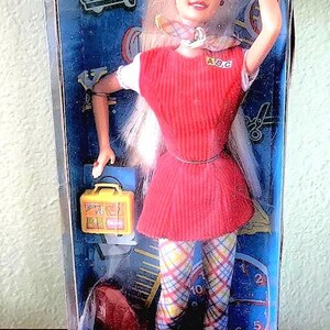 May include: A pink and blue Barbie doll box with a blonde doll wearing a red dress and plaid pants. The box says "Schooltime Fun Barbie" and has a red apple logo. The doll is holding a yellow lunchbox.