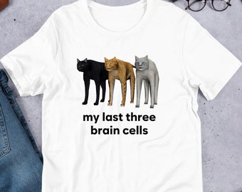 May include: White t-shirt featuring three stylized cat illustrations in black, orange, and gray. The text "my last three brain cells" is printed below the cats. The shirt is displayed on a light gray surface.