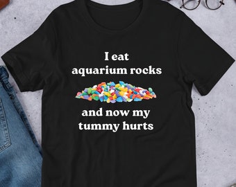 Peut inclure: T-shirt noir avec un texte blanc qui dit "Je mange des cailloux d'aquarium et maintenant j'ai mal au ventre". Un tas de cailloux d'aquarium colorés est représenté au-dessus du texte.