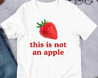Op de afbeelding: Wit T-shirt met een rode aardbei en de tekst "this is not an apple" in rood.
