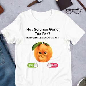 Op de afbeelding: Een wit T-shirt met een afbeelding van een sinaasappel met een orang-oetan gezicht. De tekst "Has Science Gone Too Far? Is this image real or fake?" staat op het shirt gedrukt. Er zijn twee knoppen onder de afbeelding, een met het label "Real" en de andere met het label "Fake".