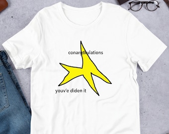 Op de afbeelding: Wit T-shirt met een gele stervormige afbeelding en de tekst "conargtkulations you've diden it".