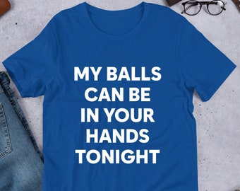 May include: Royal blue t-shirt with the white text "MY BALLS CAN BE IN YOUR HANDS TONIGHT." The shirt is displayed on a light grey surface with a pair of blue jeans, a brown wallet, a watch, glasses, and grey shoes.