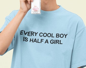 以下が含まれることがあります： 黒文字で「EVERY COOL BOY IS HALF A GIRL」とプリントされた薄い青色のTシャツ。シャツを着ている人は笑顔でジュースの箱を持っています。