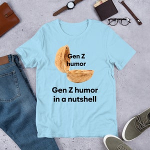 May include: Light blue t-shirt with a graphic of a walnut shell with the text "Gen Z humor" written inside the shell and the text "Gen Z humor in a nutshell" written below the shell.
