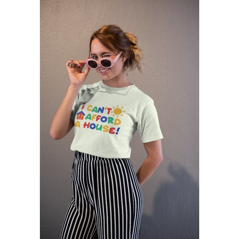 May include: A white t-shirt with a colorful graphic that says "I can't afford a house!" with a house and sun icon. The model is wearing the shirt and has pink sunglasses on.