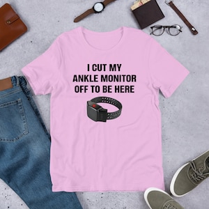 May include: Pink t-shirt with the text "I cut my ankle monitor off to be here" and a black ankle monitor with a red light on it.